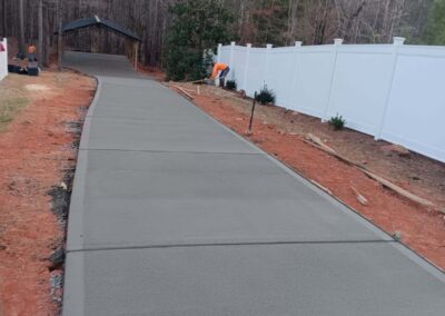 Freshly poured concrete driveway curving through a wooded area, bordered by a white fence and landscaping under construction.