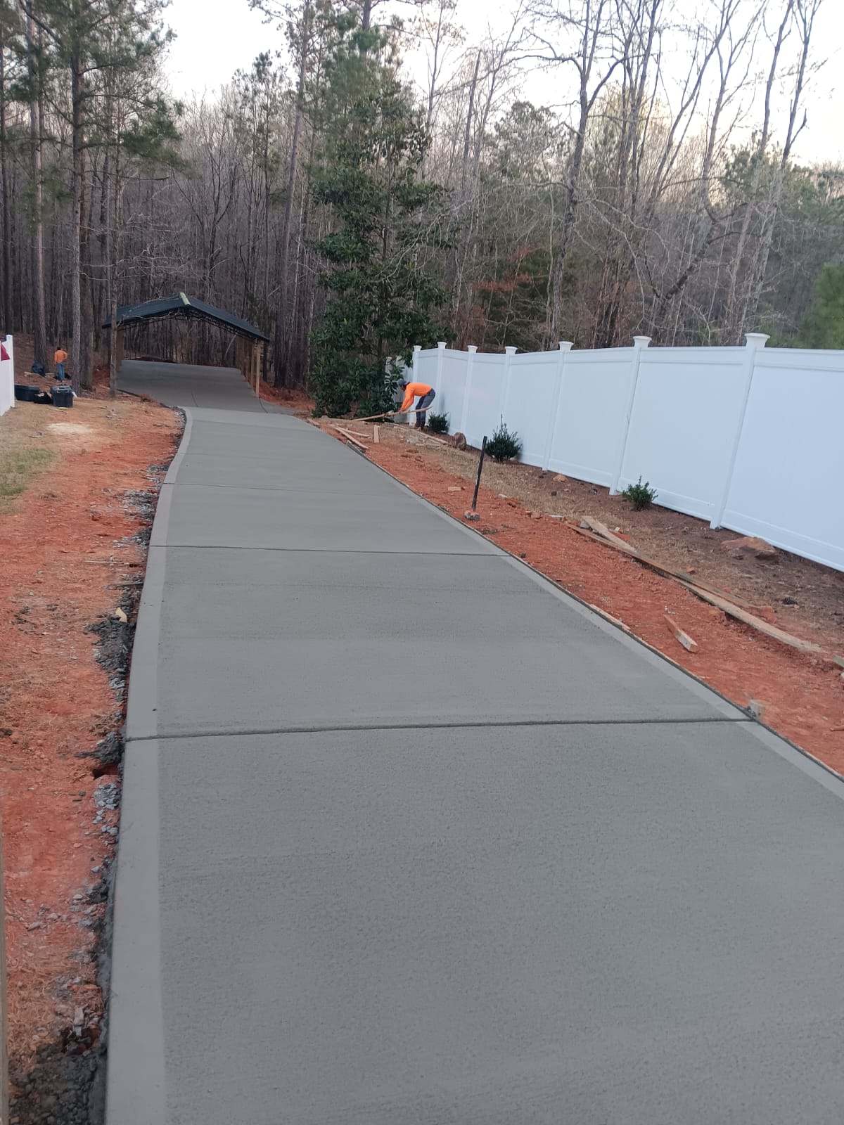 IMG-20240313-WA0003 Freshly poured concrete sidewalk curving through a wooded area, bordered by a white fence and landscaping in progress.