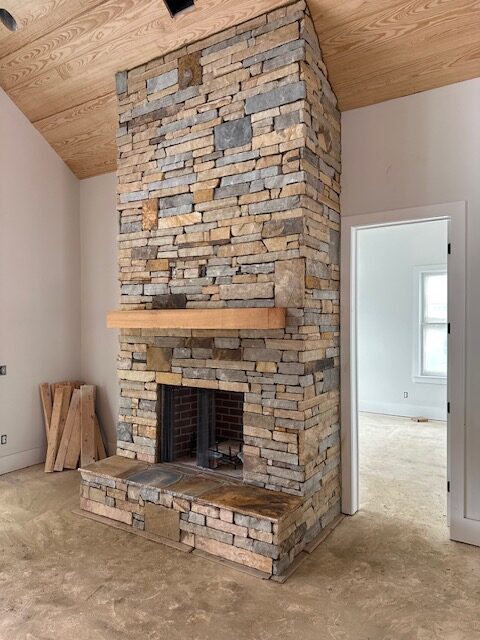 IMG_1417 Rustic floor-to-ceiling stone fireplace with a wooden mantel in a home under construction, featuring a vaulted wood-paneled ceiling.