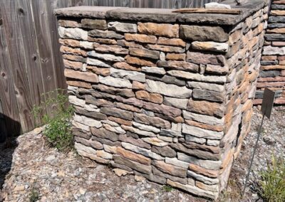 Stone wall structure, built with multi-colored stone blocks, surrounded by a gravel path. A wooden fence is visible in the background, and the stone wall is neatly stacked with a flat top edge.