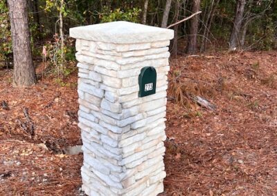 Stone mailbox pillar with a small green address plaque, situated near a wooded area with pine straw and gravel surroundings.