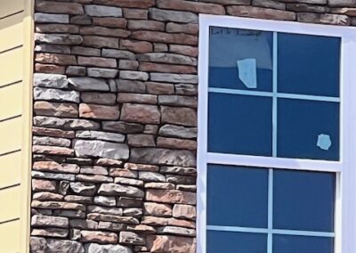 Close-up of a house exterior featuring a stone veneer facade, a white-framed window with protective stickers, and beige siding on the corner.