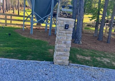 Stone pillar with a keypad entry system at the edge of a gravel driveway, with a large metal grain silo and a wooden fence in the background.