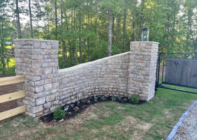 Elegant curved stone entrance wall with two pillars, decorative lanterns, a landscaped flower bed, and a wooden gate leading to a wooded property.