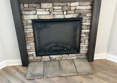 Small stone fireplace with a dark wood mantel, set against a light-colored wall with laminate wood flooring.