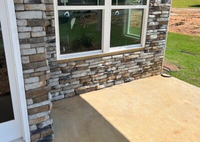 Exterior corner of a house featuring a stone veneer wall, large double-pane windows with protective stickers, and a concrete patio in bright sunlight.