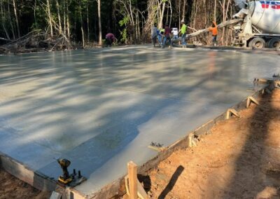 Construction workers pouring and leveling a large concrete slab foundation in a wooded area, with a cement truck on site.