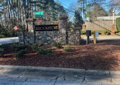 Stone entrance sign for the Brookstone community, surrounded by landscaped mulch and greenery, with a gated entrance in the background.