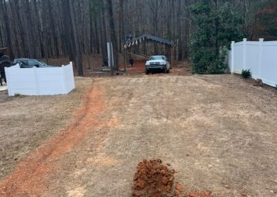 Dirt hole dug in a grassy yard leading towards a wooded area with a white fence on both sides and a metal structure in the background.
