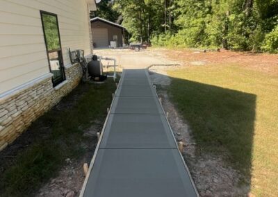 Freshly poured concrete sidewalk alongside a white building with stone veneer, leading towards another structure in a wooded area.