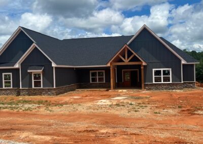 Newly constructed modern farmhouse-style home with dark gray siding, stone veneer accents, and a wooden gabled entrance, set on a lot with red soil.