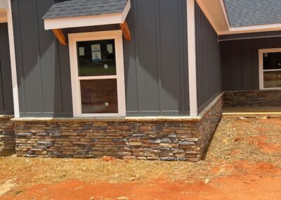 Close-up of a modern farmhouse-style home's exterior featuring dark gray board and batten siding, stone veneer foundation, and a small window awning with wood accents.
