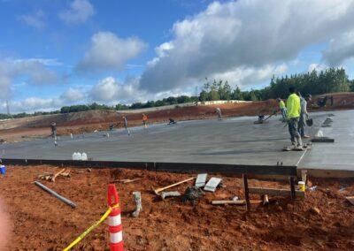Construction workers smoothing and leveling a large freshly poured concrete slab foundation on a site with red soil under a partly cloudy sky.