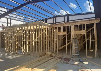 Wooden framing under construction inside a large metal building with an open roof structure, exposed steel beams, and construction materials scattered on the concrete floor.
