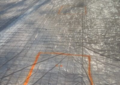Construction site with a prepared foundation covered in plastic sheeting and reinforced with wire mesh, marked with orange spray paint for designated areas.