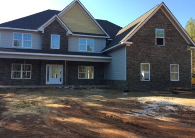 Large two-story house under construction with a combination of stone and siding exterior, multiple windows, a covered front porch, and an unfinished front yard with dirt and construction materials.