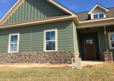 Modern green home exterior with stacked stone veneer wainscoting, white trim windows, and a covered front entrance with a wooden door under a gabled roof.