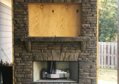Outdoor stone fireplace under construction, featuring a built-in firebox, wooden mantel, and a recessed space above for a TV or decor, framed with stacked stone veneer.