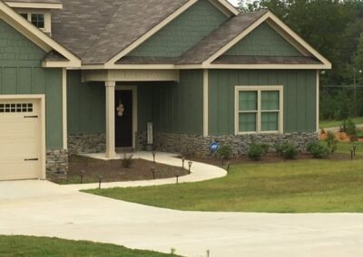 Modern single-story home with green siding and stone accents, featuring a curved concrete walkway leading to the front entrance, a landscaped yard, and a driveway.