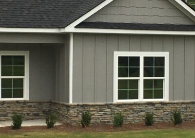 Close-up of a modern home exterior with gray board and batten siding, white trim, and a stone veneer base, complemented by neatly arranged landscaping.