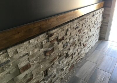 Close-up view of a modern accent wall featuring stacked stone veneer in shades of white, gray, and brown. A wooden trim runs along the top of the stone, complementing the textured design. The floor is covered with large rectangular gray tiles, enhancing the contemporary aesthetic.
