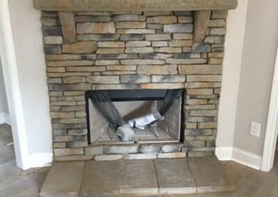 Rustic stone fireplace with a thick stone mantel in a modern home. The hearth is made of large stone slabs, and the firebox is covered with protective plastic. The surrounding walls are painted light gray, and the floor is covered with wood-look laminate flooring.