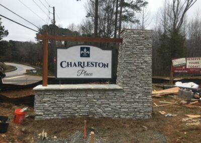 Entrance sign for 'Charleston Place' featuring a white and blue sign mounted on a stone veneer base with a wooden beam accent, situated near a road with trees and construction materials in the background.