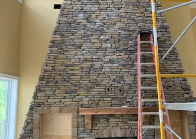 Large floor-to-ceiling stone fireplace under construction in a high-ceiling living room. A wooden mantel is installed, with built-in shelving areas on either side. A ladder and scaffolding are set up for ongoing work, with construction materials and tools scattered around.