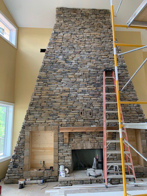 Large floor-to-ceiling stone fireplace under construction, featuring a wooden mantel, built-in shelving, scaffolding, and a ladder in a high-ceiling living room.