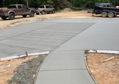 Freshly poured concrete driveway and walkway in a construction site, with a curve leading to a large flat area. Trucks and a trailer are parked nearby, and the surrounding area is still under construction, with trees visible in the background.