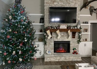 Cozy living room decorated for Christmas, featuring a large stone fireplace with a TV mounted above it, stockings hanging from the mantle, and a glowing electric fireplace below. A festive Christmas tree with lights and ornaments stands nearby. The room is warm and inviting, with a neutral-colored rug and furniture.