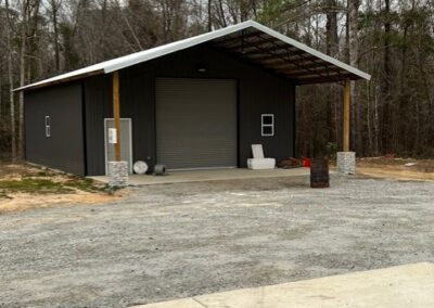 Large black metal storage building with a slanted roof, featuring a roll-up garage door, small windows, and stone pillar accents at the corners. A gravel driveway leads up to the building, with trees in the background.