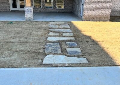 Pathway made of large, natural stone slabs laid across a grassy area in front of a brick building with multiple windows. The grass is not fully grown, and the concrete sidewalk leads to the stone path.