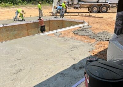 Construction workers pouring and smoothing concrete around an unfinished in-ground pool, with a USA-branded cement truck in the background on a dirt lot.