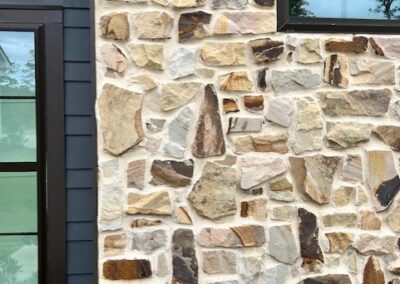 Close-up view of a natural stone veneer exterior wall with various shades of beige, brown, and tan stones, adjacent to a dark-colored siding and black-framed windows.