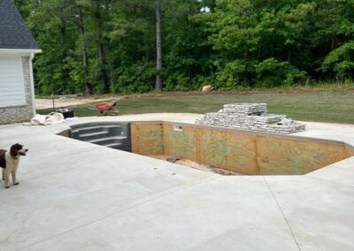 Backyard construction site with an unfinished in-ground pool surrounded by a concrete patio, stacked stone materials, and a dog standing nearby.